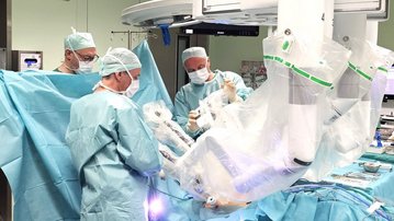 Ein chirurgisches Team in einem Operationssaal führt einen Eingriff durch. Die Ärzte tragen Schutzkleidung und Masken, während sie sich auf den Patienten konzentrieren, der von blauen Tüchern umgeben ist.