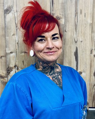 Eine Person mit roten Haaren und sichtbaren Tattoos trägt ein blaues Oberteil. Sie hat große Ohrringe und ein Piercing in der Nase. Der Hintergrund besteht aus Holzpaneelen.