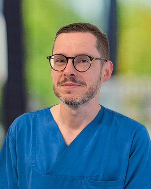Mann in blauer Arbeitskleidung mit Brille, steht lächelnd vor einem unscharfen Hintergrund.