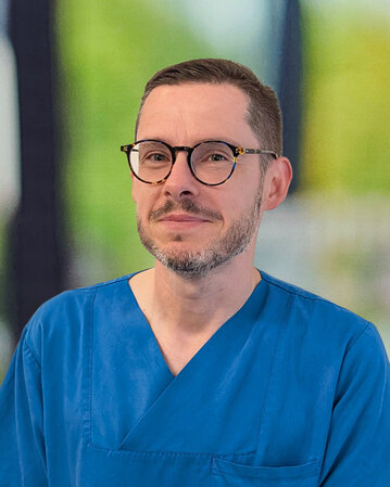Mann in blauer Arbeitskleidung mit Brille, steht lächelnd vor einem unscharfen Hintergrund.