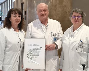 Drei medizinische Fachkräfte in weißen Kitteln stehen in einem Flur und halten ein Zertifikat oder eine Auszeichnung in der Hand.