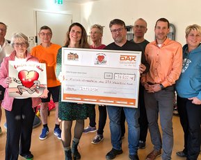 Eine Gruppe von Menschen hält einen großen Scheck und ein Herz-Symbol. Sie stehen zusammen in einem Raum, lächelnd und feierlich.