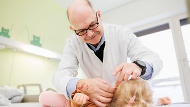 Ein Arzt untersucht ein Kleinkind, das auf einer Liege liegt. Der Arzt trägt eine Brille und einen weißen Kittel.