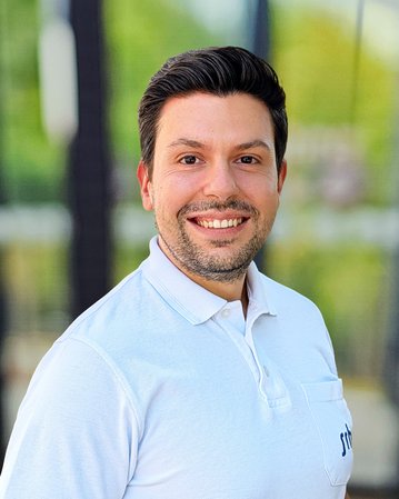 Ein Mann mit dunklen Haaren und Bart lächelt in einem weißen Poloshirt, vor einer unscharfen, grünen Hintergrundlandschaft.