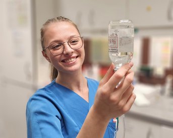 Eine Krankenschwester in blauer Uniform hält einen Tropf mit Flüssigkeit und lächelt dabei.