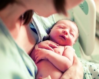 Eine Mutter hält ein neugeborenes Baby in ihren Armen, das entspannt und schlafend aussieht, während sie in einem Krankenhausbett sitzen.