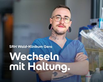 Ein Mann mit Tattoos und großen Ohrsteckern trägt einen blauen Krankenhauskittel und steht mit verschränkten Armen in einer Klinik.