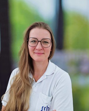 Eine Frau mit langen, blonden Haaren und einer weißen Bluse steht mit verschränkten Armen vor einem unscharfen, grünen Hintergrund. Sie trägt eine Brille und lächelt leicht.