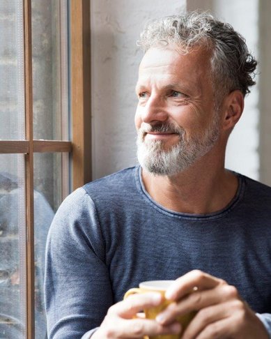 Ein Mann mit grauem Bart und lockigem Haar sitzt am Fenster und hält eine Tasse in der Hand. Er blickt nachdenklich nach draußen.