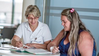 Chefärztin Prof. Stroh und eine Patientin sitzen an einem Tisch und besprechen Informationen zum Thema Adipositas. Die Ärztin zeigt auf eine Seite, während die Patientin aufmerksam zuhört.