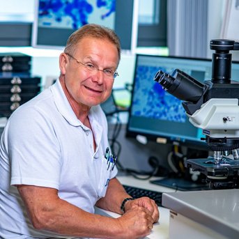 Chefarzt Prof. Uwe Will, Klinik für Gastroenterologie, Hepatologie, Diabetologie und Allgemeine Innere Medizin Ein Arzt sitzt an einem Schreibtisch und schaut in ein Mikroskop. Im Hintergrund sind Computerbildschirme mit wissenschaftlichen Daten sichtbar.