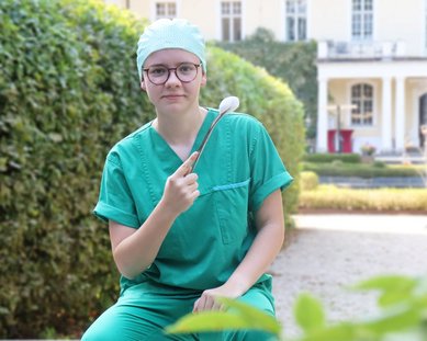 Eine Person - Auszubildende Mailin - in grüner OP-Kleidung hält einen Tupfer und sitzt im Schweinsburger Park, umgeben von Pflanzen und einem gepflegten Garten.
