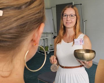 Eine Frau mit langen, blonden Haaren hält eine Klangschale und einen Schlägel, während sie einer anderen Person gegenübersteht.