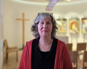 Eine Frau mit grauen Haaren und Brille steht in einem Raum mit einem Kreuz und mehreren Bildern im Hintergrund.
