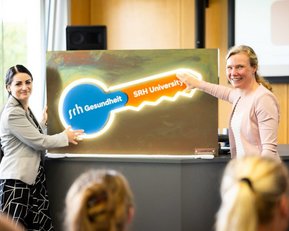 Zwei Frauen präsentieren einen großen Schlüssel mit dem Logo der SRH Universität während einer Veranstaltung.