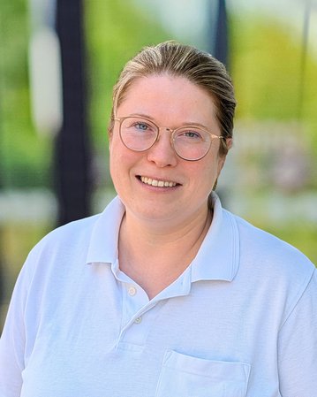 Eine lächelnde Frau mit Brille trägt ein weißes Polohemd mit dem Logo "srh". Der Hintergrund zeigt unscharfe grüne Bäume.