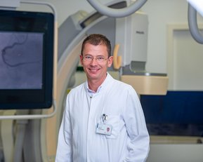 Ein Arzt in einem weißen Kittel steht in einem modernen medizinischen Raum mit einem großen Bildschirm im Hintergrund.