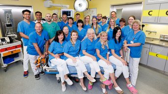 Eine Gruppe von 20 Personen in blauen Uniformen steht in einem Krankenhausraum, umgeben von medizinischer Ausrüstung und Geräten.