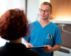 Ein Arzt in blauer Uniform spricht mit einer Patientin, während er ein Tablet in der Hand hält. Die Patientin hat lockige, rote Haare.