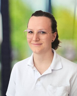 Eine Person mit Brille trägt ein weißes Polohemd mit einem Logo auf der Brust. Der Hintergrund zeigt unscharfe grüne und blaue Elemente.