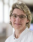 Eine Person mit blonden, lockigen Haaren und einer weißen Arztkittel steht in einem hellen, modernen Innenraum. Sie trägt eine Brille und lächelt leicht.