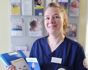 Eine Person in blauer Arbeitskleidung hält ein Informationsblatt und eine Mappe in der Hand, vor einem Informationsbereich mit Broschüren.