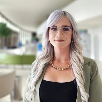 Eine Person mit langen, blonden Haaren und Brille steht in einem modernen Innenraum. Sie trägt ein schwarzes Oberteil und eine grüne Jacke. Im Hintergrund sind unscharfe Figuren und Pflanzen zu sehen.