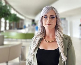 Eine Person mit langen, blonden Haaren und Brille steht in einem modernen Innenraum. Sie trägt ein schwarzes Oberteil und eine grüne Jacke. Im Hintergrund sind unscharfe Figuren und Pflanzen zu sehen.