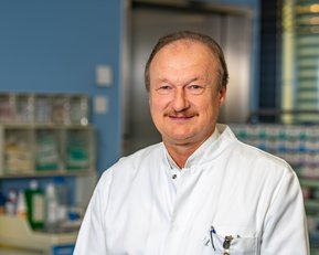 Ein Arzt in einem weißen Kittel steht in einem medizinischen Raum mit Regalen voller medizinischer Utensilien im Hintergrund.