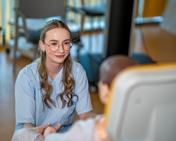 Eine Pflegekraft spricht mit einem Patienten, der im Bett sitzt. Die Pflegekraft trägt ein hellblaues Outfit und hat lange, lockige Haare.