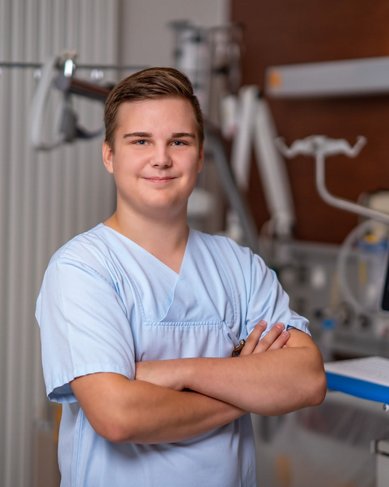 Ein junger Mann in einem hellblauen medizinischen Outfit steht mit verschränkten Armen vor einem modernen medizinischen Bildschirm in einer Klinik.